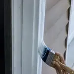 Painter brushing dark blue paint onto exterior window trim on a home with lap siding.