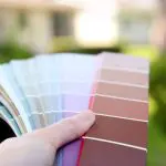 Hand holding a fan of exterior paint color swatches outdoors with a blurred home and green landscaping in the background.