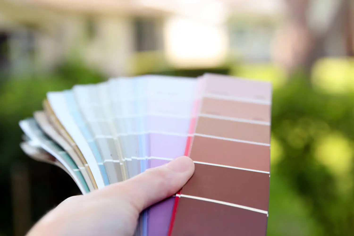 Hand holding a fan of exterior paint color swatches outdoors with a blurred home and green landscaping in the background.