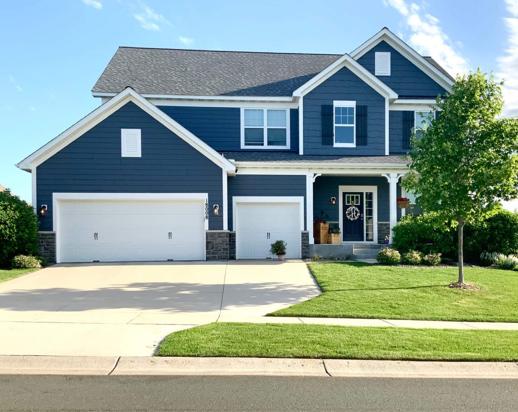Elegant two-story blue house with white trim and a three-car garage, showcasing professional exterior house painting in Ramsey MN with a modern, polished finish.