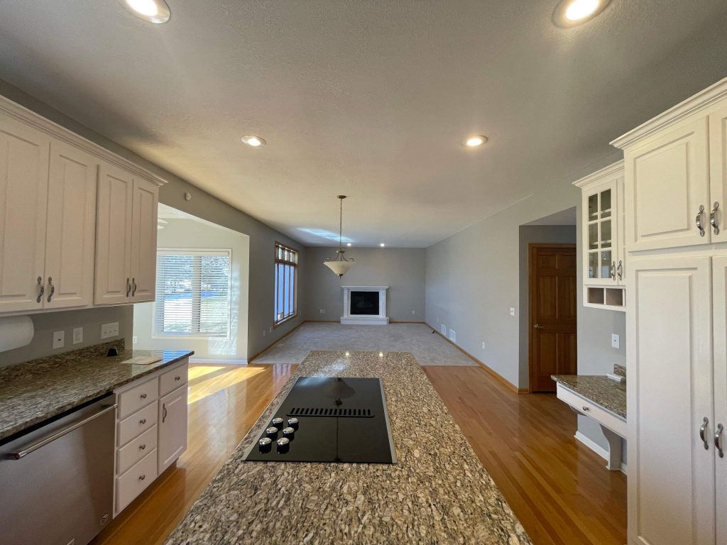 Interior Painters in Ramsey, MN: An open-concept house interior, showing a kitchen with white cabinets and granite countertops, a central island with a stovetop, wood floors, and a living room with a fireplace, featuring freshly painted gray walls.