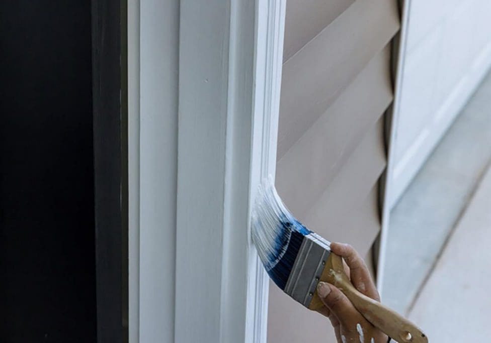Painter brushing dark blue paint onto exterior window trim on a home with lap siding.