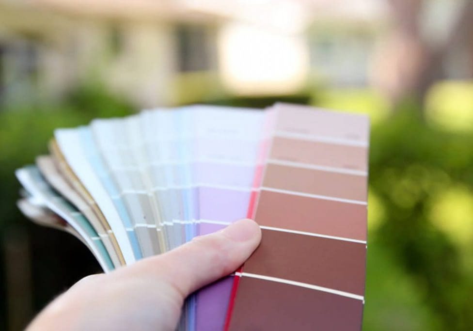 Hand holding a fan of exterior paint color swatches outdoors with a blurred home and green landscaping in the background.