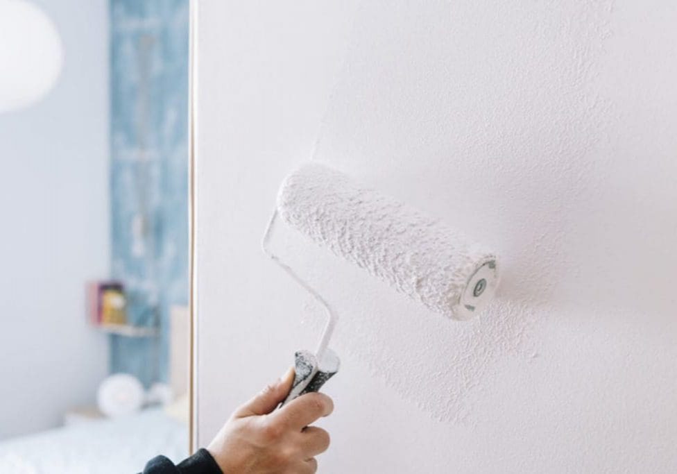 Painter rolling white paint onto an interior wall with blue tile visible in the background.